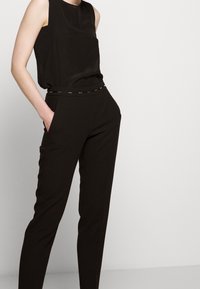 Černý bezrukávový top v kombinaci s černými zúženými kalhotami. Kalhoty mají tenký opasek s kovovými akcenty a boční kapsy. Hladká textura.