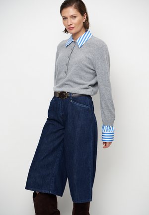Femme portant un pull gris par-dessus une chemise rayée bleue et blanche, un pantalon large en jean foncé et des bottes marron, debout devant un fond blanc.