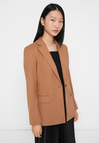 Blazer marrone con una vestibilità su misura, chiusura a un bottone e revers, indossato su un top nero, abbinato a una gonna plissettata.