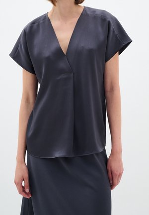 Nainen pukeutuneena tummanharmaaseen, teksturoituun V-kaula-aukkoiseen lyhythihaiseen puseroon ja vastaavaan hameeseen vaaleaa taustaa vasten, keskivartalon näkymä.