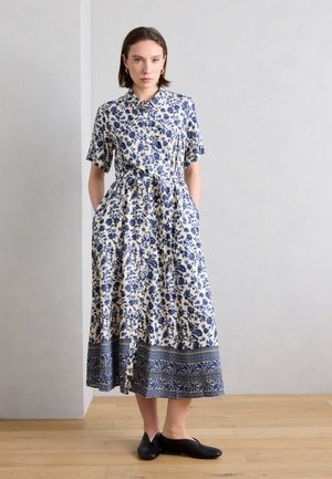 Kvinde stående iført en hvid midi-kjole med blå blomsterprint, korte ærmer, krave, taljebånd og sorte flade sko på trægulv.