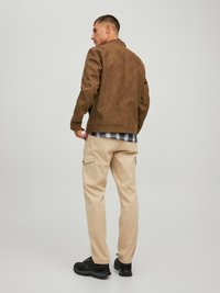 Giacca in suede marrone con una trama liscia, pantaloni cargo beige e sneakers nere. L'outfit presenta una camicia a quadri visibile sotto.