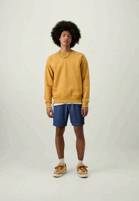 Νεαρός άντρας με σγουρά μαλλιά, φορώντας ένα κίτρινο φούτερ, μπλε τζιν σόρτς, λευκές κάλτσες και κίτρινες αθλητικές sneakers, στέκεται μπροστά από μια απλή φόντο.