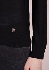 Svart stickad tröja med långa ärmar, med en figurnära ribbad nederkant och en silvrig logotypdetalj nära den nedre kanten.