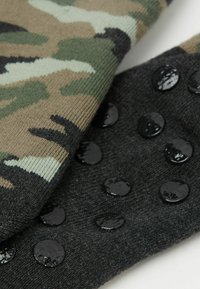 Dos calcetines: uno con un patrón de camuflaje en tonos de verde y marrón, y el otro en negro con puntos circulares de silicona brillante para agarre.
