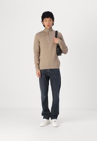 Pulover bej cu fermoar la gât, cu manșete și tiv cu textură ribbed, asortat cu blugi de un albastru închis și adidași albi, ținând o mică geantă neagră.
