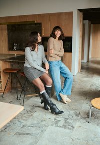 Deux femmes assises sur des tabourets de bar dans une cuisine moderne. L'une porte un sweat à fermeture éclair gris et une jupe à carreaux, l'autre un haut en tricot beige et un jean bleu. Toutes deux portent des bottes montantes.
