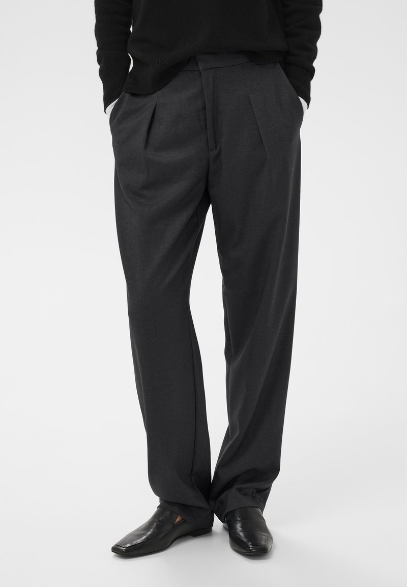 Sive plisirane hlače napravljene od lagane tkanine, s opuštenim krojem. Usklađene s sjajnim crnim cipelama.