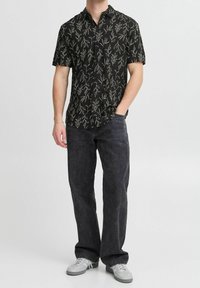 Korte mouwen zwart shirt met witte bladerenprint, gecombineerd met donkere denim broek. Model draagt lichtgrijze sneakers. Casual fit en stijl.