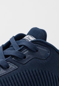 Sportovní bota námořnické modré barvy s texturovaným pleteným svrškem, plochými tkaničkami a lesklou logo cedulkou na jazyku. Zvýrazněná hladkými detaily.