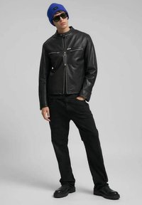 Zwarte leren jas met opstaande kraag, ritssluiting op borstzakken, gecombineerd met zwarte broek en een blauwe muts. Model draagt een zonnebril.