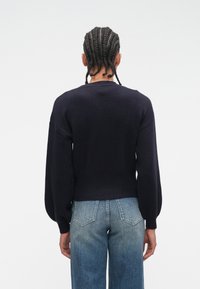 Person with braided hair wearing a dark long-sleeve sweater and blue denim jeans shown from the back against a plain background.