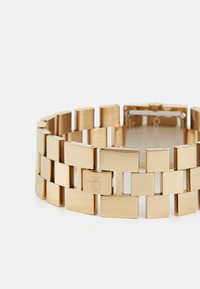 Reloj de pulsera de metal dorado con eslabones cuadrados. Cuenta con un cierre de broche y un logotipo grabado en un eslabón rectangular. Acabado pulido.