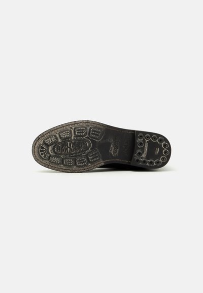 Suela de goma negra de un zapato que presenta un patrón de agarre texturizado, un logo y acentos circulares. La superficie muestra un aspecto desgastado.