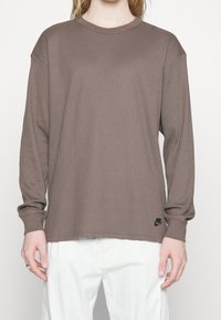 Hnědé tričko s dlouhým rukávem z žebrované látky, s kulatým výstřihem a černým logem Nike na levém lemu. Nosit se bílé kalhoty.