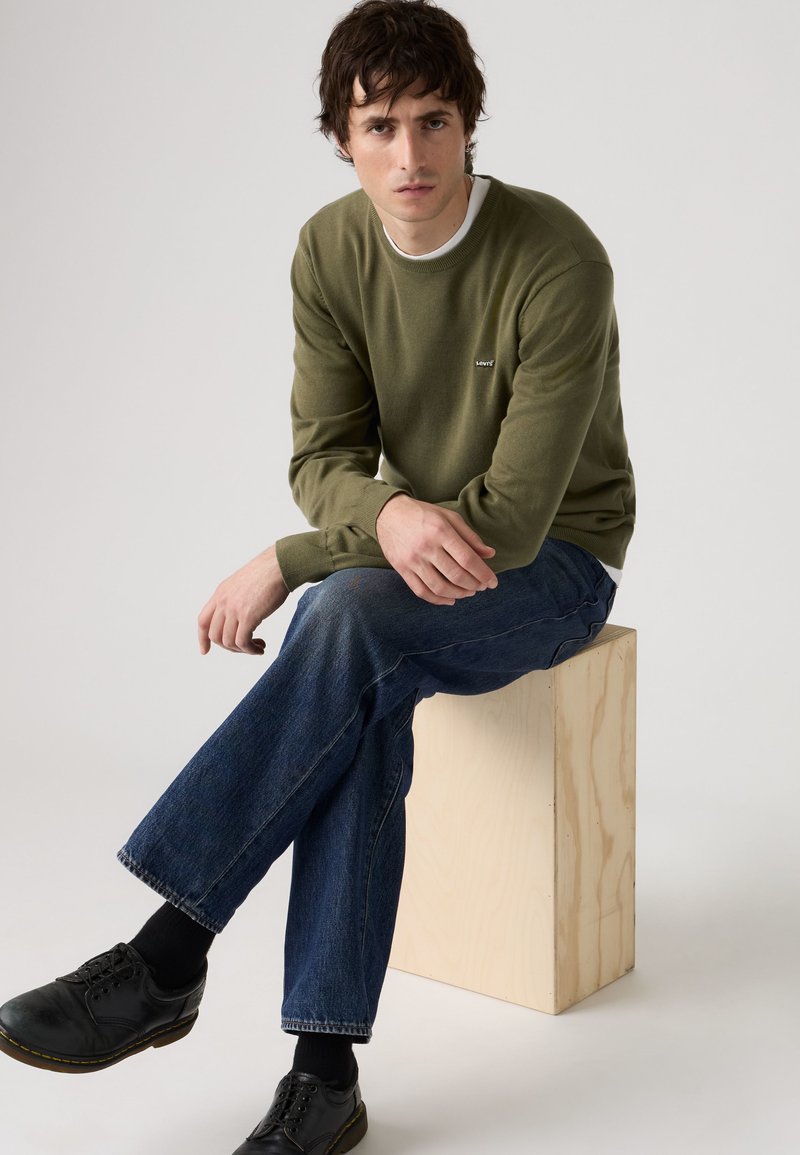 Man with dark hair wearing an olive green sweater and blue jeans sitting on a wooden block against a plain background.