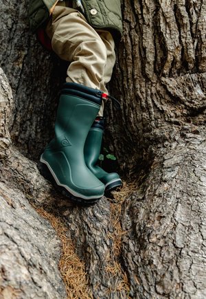 Kind trägt grüne Gummistiefel und beige Hose steht in der Höhlung eines großen Baumstamms im Freien.