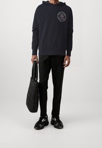 Námořnická mikina s kapucí a grafickým logem na hrudi, spárovaná s černými kalhotami a prošívanou černou taškou. Černé sportovní tenisky dodávají vzhledu finální úpravu.