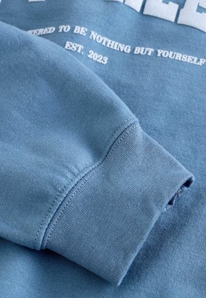Blue cotton sweatshirt with a soft texture. Features a ribbed cuff and printed text in white, including "EST. 2023" and additional motivational phrases.