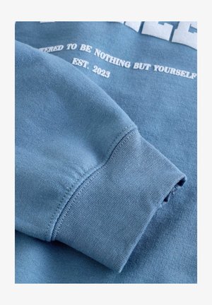 Felpa in cotone blu con una texture morbida. Presenta un polsino a costine e testo stampato in bianco, inclusi "EST. 2023" e ulteriori frasi motivazionali.