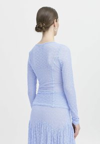 Top lungo a maniche in pizzo lilla con un design aderente, caratterizzato da intricati motivi floreali e un collo a giro, abbinato a una gonna coordinata.