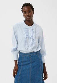 Donna con capelli intrecciati che indossa una blusa azzurra con volant e una gonna di jeans blu a vita alta, su uno sfondo bianco uniforme.