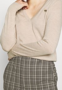 Pull en maille beige avec un col en V, des manches longues et une broche florale sur le côté gauche, associé à une jupe écossaise taille haute.