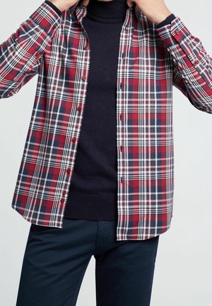 Man knoopt een rode, marineblauwe en witte geruite blouse over een zwarte coltrui met marineblauwe broek tegen een effen achtergrond.