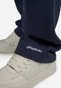 Marineblaue gerippte Sweatpants mit umgeschlagenem Saum, die weiße "Degador"-Stickerei zeigen, kombiniert mit cremefarbenen Sneakers mit strukturierten Details.