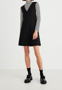 Robe noire sans manches à encolure en V, portée par-dessus un haut à manches longues rayé noir et blanc. Comprend des chaussures à plateforme noires épaisses et des socquettes.