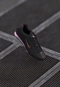 Crne i smeđe Nike tenisice s ru�žičastom potplatom lebde iznad metalne rešetkaste podloge s preklapajućim dijagonalnim linijama.