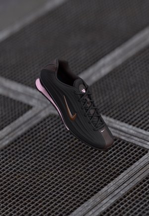 Sapatilha Nike preta e castanha com sola rosa flutuando acima de um chão de grelha metálica com linhas diagonais interseccionadas.