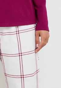 Haut violet à manches longues associé à un pantalon à carreaux blancs avec des lignes rouges. Texture lisse avec des manches ajustées, montrant une main avec des bagues.