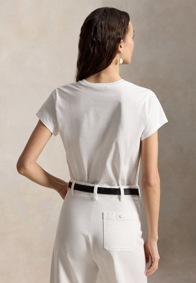 Γυναίκα με μακριά σκούρα μαλλιά, ντυμένη με λευκό πουκάμισο με κοντά μανίκια που είναι φαγωμένο μέσα σε λευκό παντελόνι με μαύρη ζώνη, στέκεται με το ένα χέρι στη μέση.