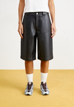Sorte skinnshorts med store sidelommer, en litt løs passform, og knappelukking, båret med hvite sokker og fargerike joggesko.