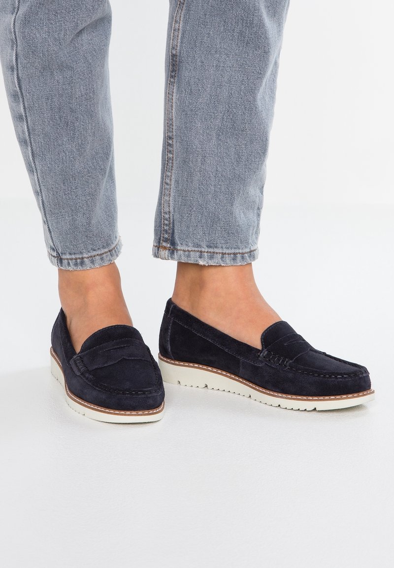 Mocassins en daim bleu marine avec un détail cousu et une semelle en caoutchouc crème. Portés avec un jean gris ourlé, mettant en valeur un style décontracté.