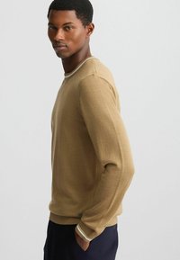 Hellbraunes Crewneck-Pullover mit langen Ärmeln, gerippten Bündchen und einem kontrastierenden weißen Streifen am Halsauschnitt. Eine glatte, weiche Textur.