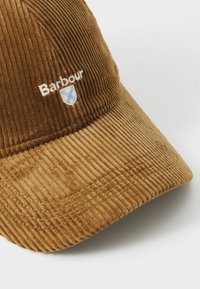 Bruine corduroy baseballcap met een gebogen rand en een geborduurd 'Barbour'-logo en schild op het voorpaneel tegen een witte achtergrond.