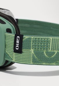 Žalios slidinėjimo akiniu su raštuotu dirželiu. Dirželis pagamintas iš tekstūruoto žalio audinio ir turi juodą GIRO logotipo etikėtę. Lygus plastikinis rėmas.