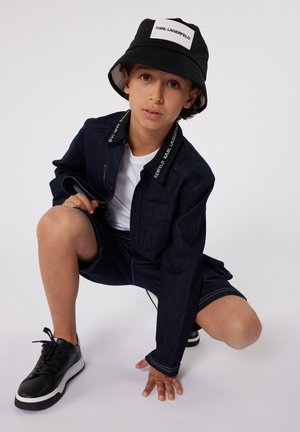 Veste en denim bleu avec des accents noirs, t-shirt blanc, shorts sombres et baskets noires. Le chapeau seau présente une étiquette logo et un doublure noire.