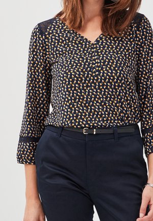 Femme portant un chemisier bleu marine avec de petits motifs géométriques orange et blanc et un pantalon bleu marine avec une ceinture noire.