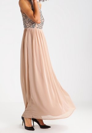 Femme portant une robe sans manches beige avec un corsage orné et une jupe fluide, assortie à des chaussures à talons hauts noires.