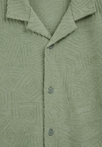 Chemise vert clair texturée avec des motifs géométriques en relief et trois boutons vert foncé sur le devant, avec un col plié.