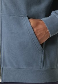 Sudadera azul con un bolsillo frontal. Tela suave, puños y dobladillo acanalados. Cierre con cremallera y costuras planas, que muestran una textura suave.