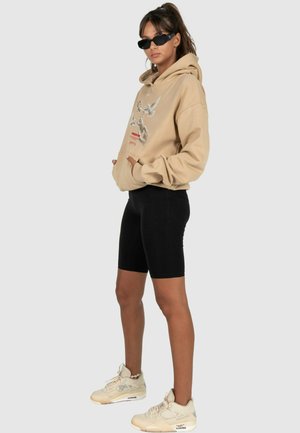 Beige Kapuzenpullover mit grafischem Druck, schwarze enganliegende Shorts und beige Sneakers. Merkmale sind das übergroße Design, Taschen und Sonnenbrille.