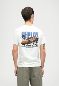 Vīrietis valkā baltu T-kreklu ar oranžā sacīkšu auto, palmu kokiem, šaha karogu un uzrakstu "REPLAY" aizmugurē.