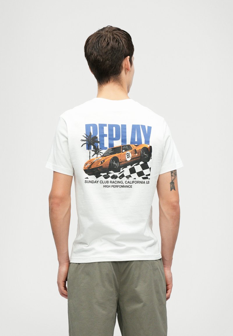 Vīrietis valkā baltu T-kreklu ar oranžā sacīkšu auto, palmu kokiem, šaha karogu un uzrakstu "REPLAY" aizmugurē.