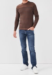 Braun gestreifte Pullover mit rundem Halsausschnitt und Knopfakzenten an der Schulter, kombiniert mit blauen Jeans und weißen Turnschuhen.