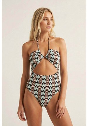 Mujer rubia con traje de baño tipo halter estampado con diseño de zigzag y rayas, posando contra un fondo liso y claro.