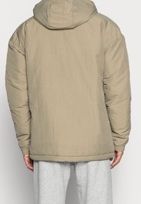 Beige vadderad jacka med huva, med en slät textur och elastiska ärmmuddar. Designen är avslappnad passform och sträcker sig till höfterna.